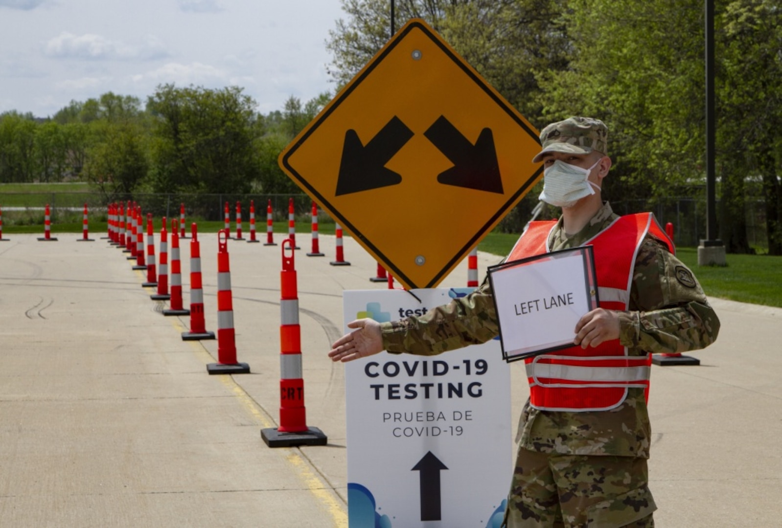 An Army PFC wearing a uniform, reflective vest and mask directs traffic with his arm extended in front of a directional sign for COVID-19 testing site.