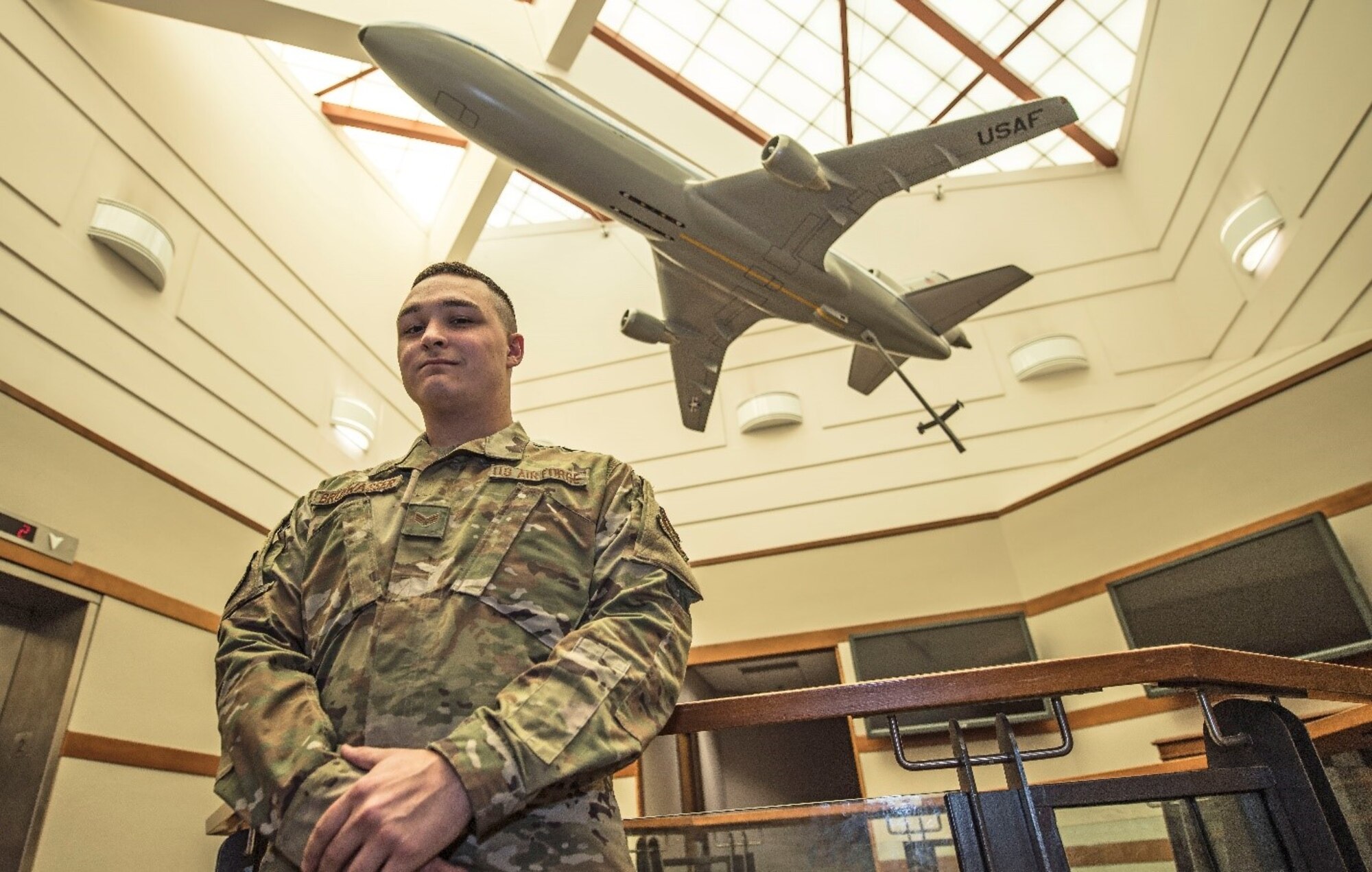 A man in a military uniform poses inside a building while a model of a KC-10 Extender is suspended above him.