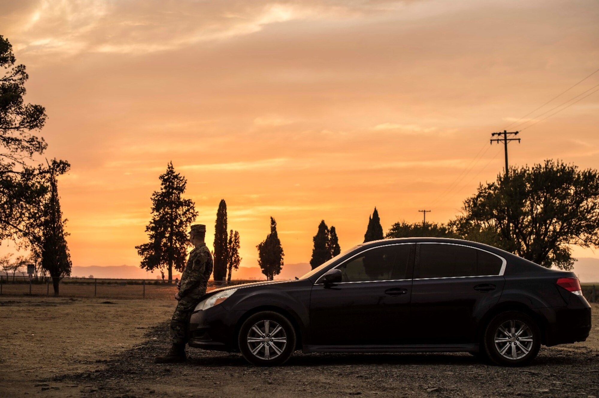 A man in a military uniform sits against his car at sunset