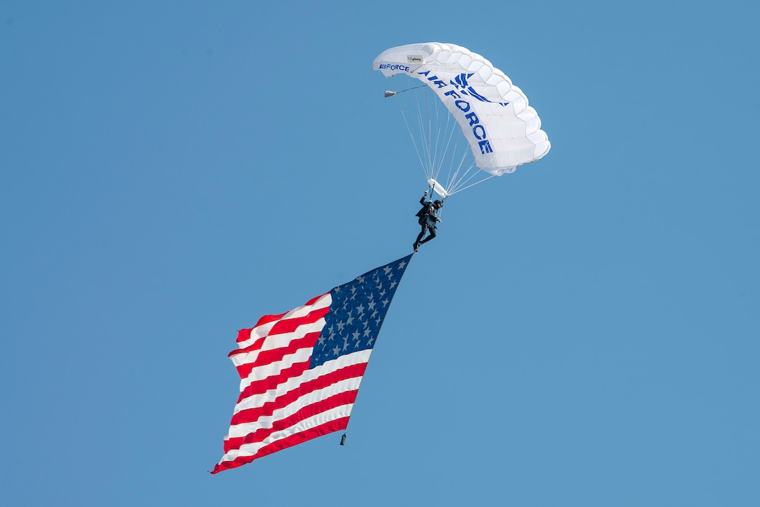 A parachutist with a white parachute with the Air Force logo descends in blue sky with an American flag.
