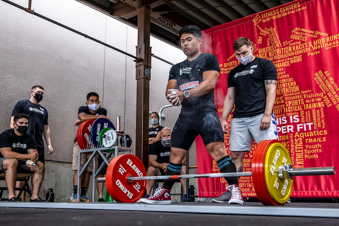A Marine in gym clothes stands with his legs apart and white powder on his hands in front of a barbell as fellow Marines in face masks watch.