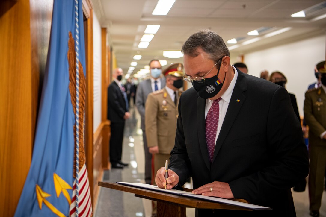 A man signs a book as people wait in a hallway behind him.