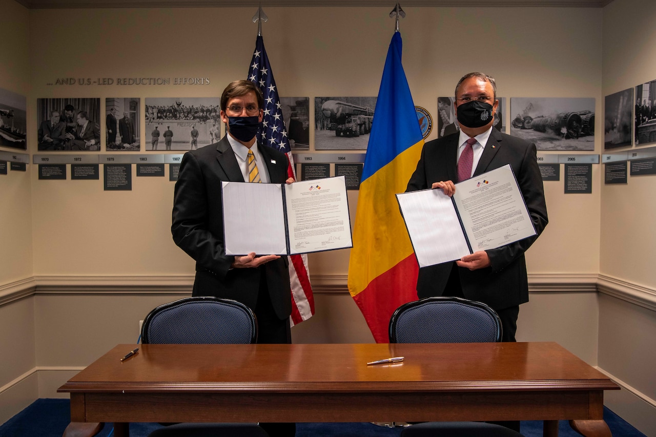 Two men stand in front of flags holding documents.