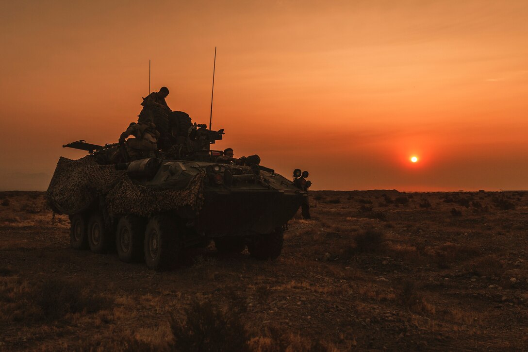 Marines, shown in silhouette, work atop a military vehicle with an orange sky as the backdrop.