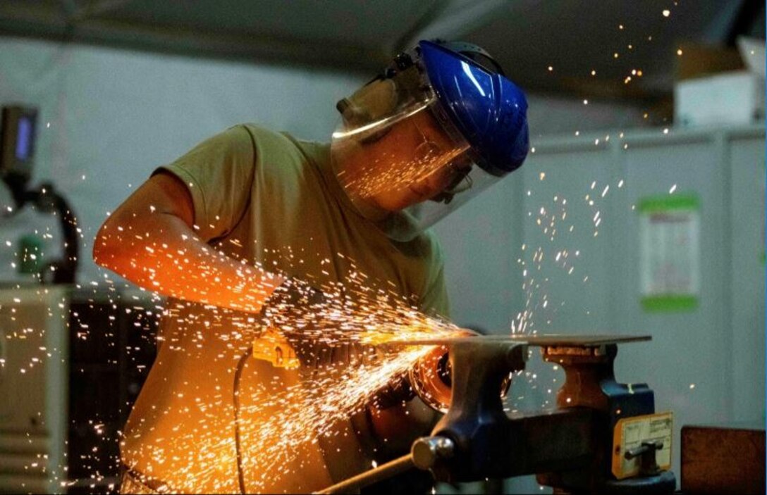 An airman wearing protective gear grinds metal as sparks fly.