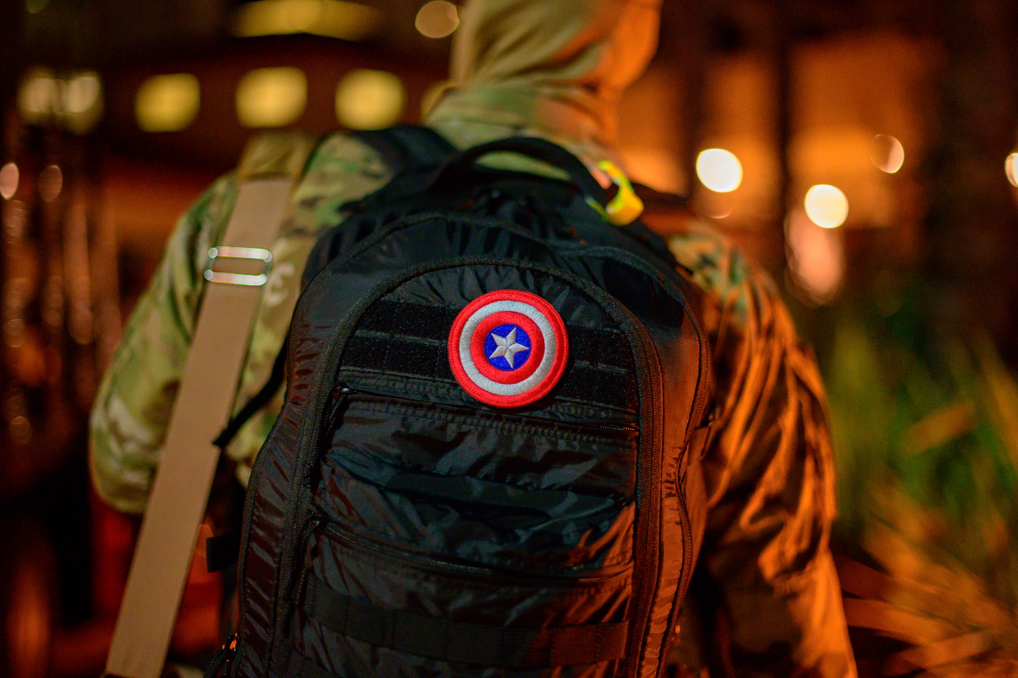 An Airman stands in line to board a bus while wearing a black backpack with a Captain America patch in the center of his backpack.