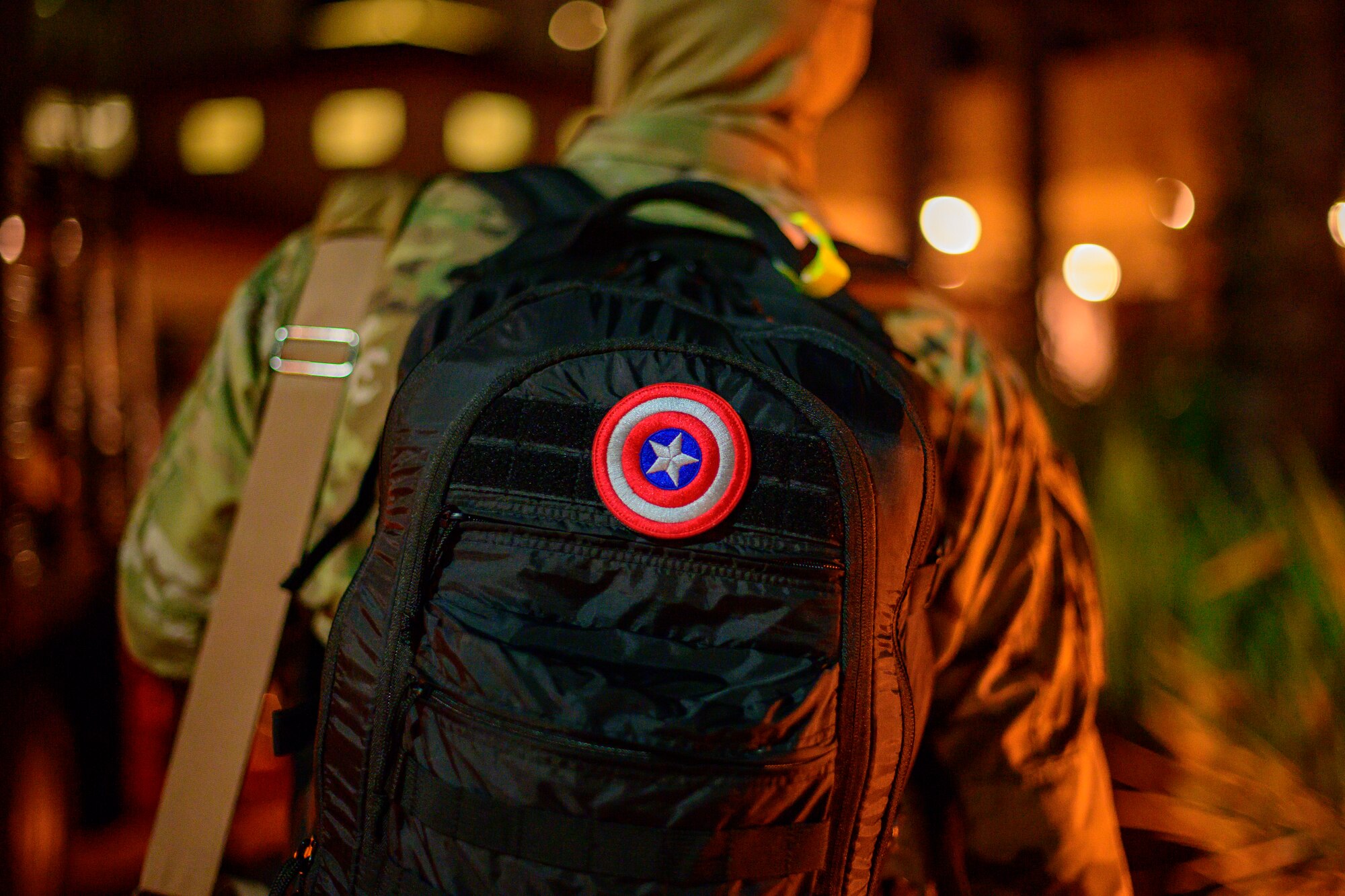 An Airman stands in line to board a bus while wearing a black backpack with a Captain America patch in the center of his backpack.