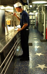 Mess Specialist 2nd Class Eric Oliver makes final menu preparations on the ship's mess decks.  At his feet are white stars in an area renamed as the "Hall of Heroes."