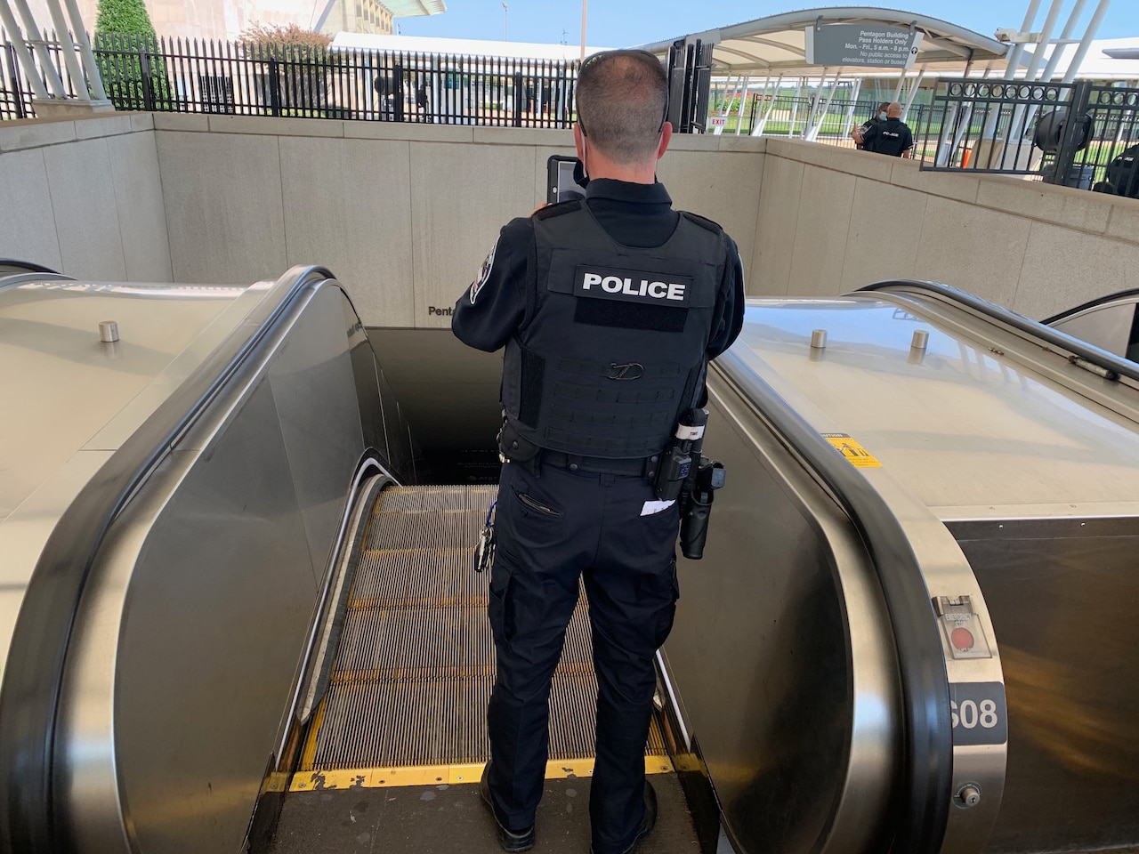 A Pentagon police, shown from behind, stands and holds up a tablet at the top of an escalator.