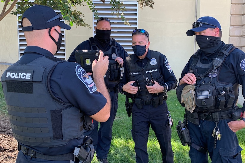 Three police officers wearing face masks stand and look toward a tablet a fellow officer holds in front of them outside.