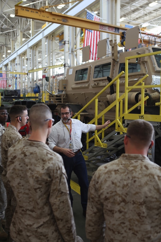 Several men in military uniforms listen as a man in civilian clothes speaks.  A military vehicle is on an assembly line behind the men.