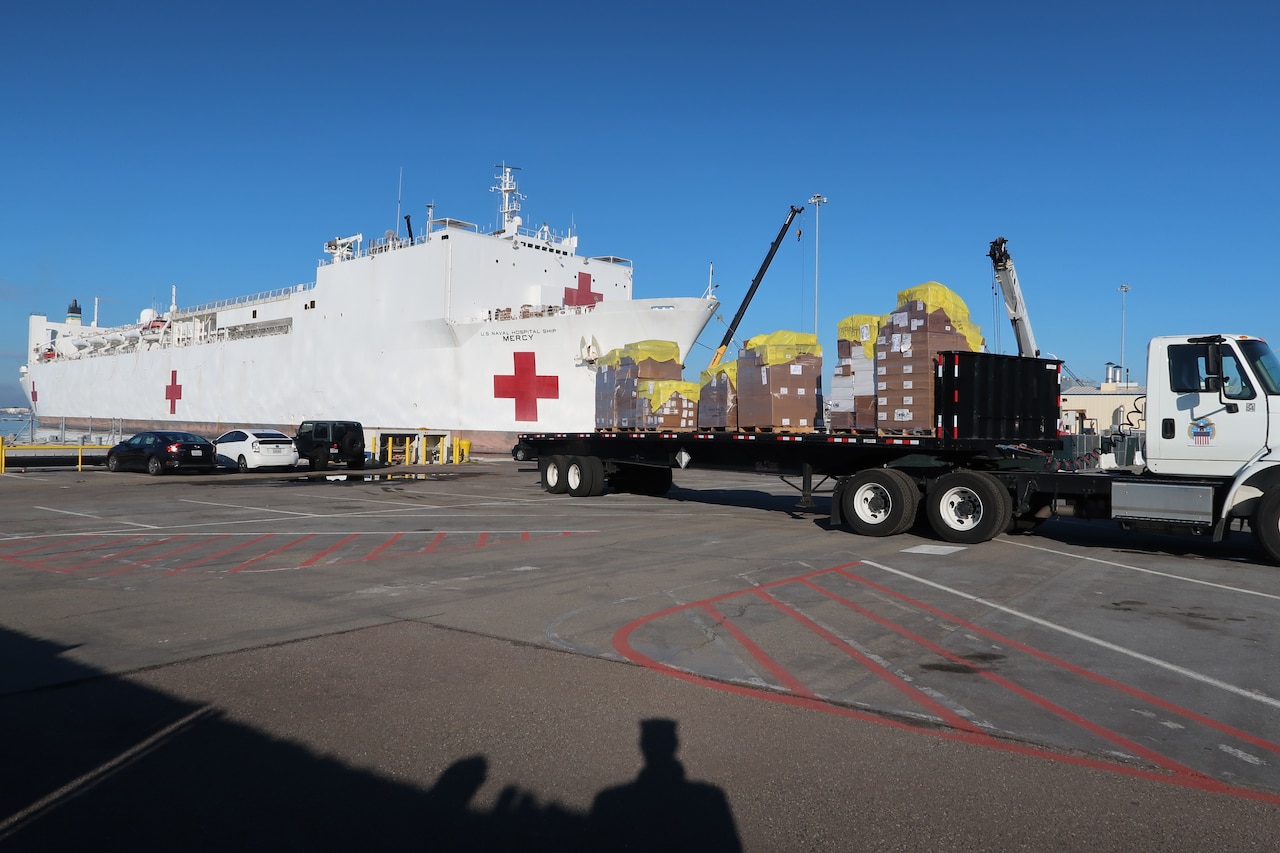 A truck loaded with pallets waits on a concrete lot. Nearby, a naval vessel bearing a red cross is docked.