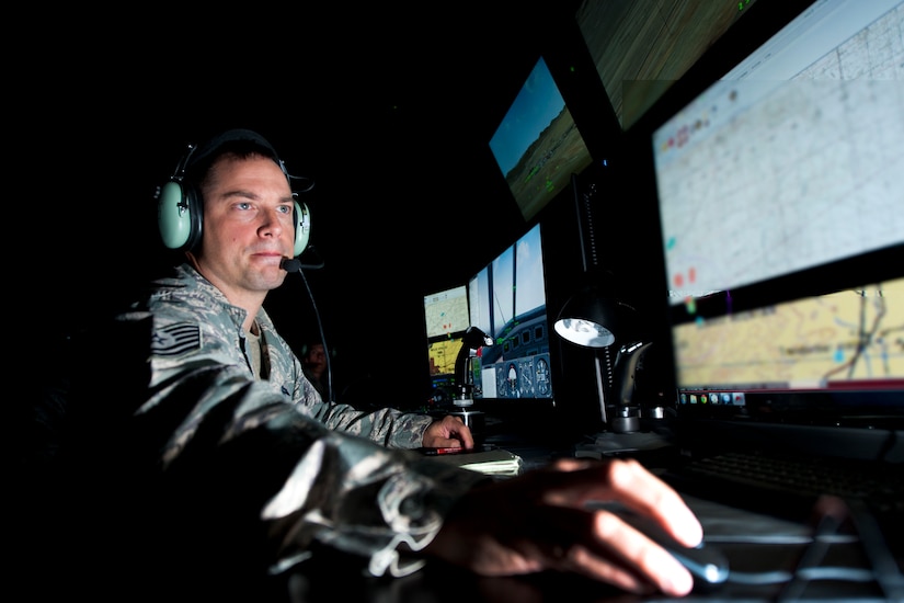 A man in military uniform wears a headset in a room lit only by computer screens.