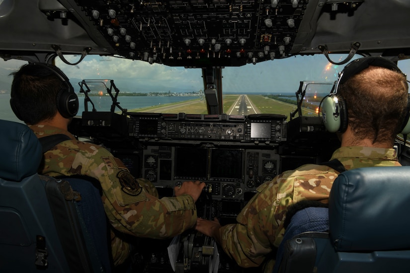 In a view from the back of a cockpit, two pilots look ahead at an airport runway.