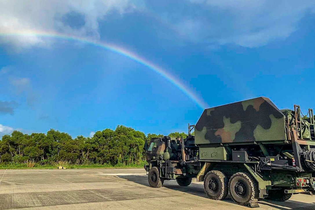 A military vehicle sits in a parking lot as a rainbow shines in the sky.