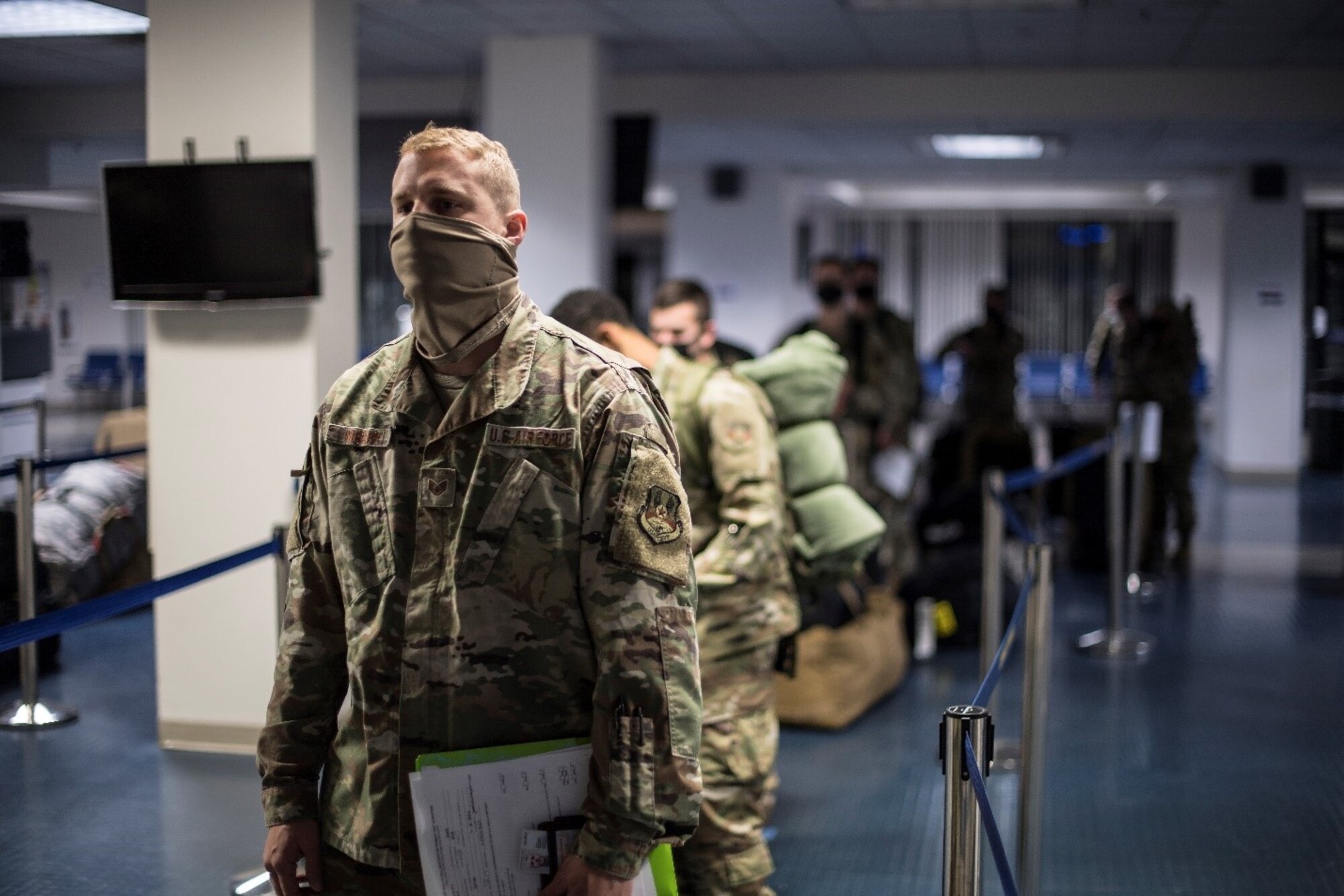 Men and women in military uniforms and face masks wait in a line inside airport-like surroundings