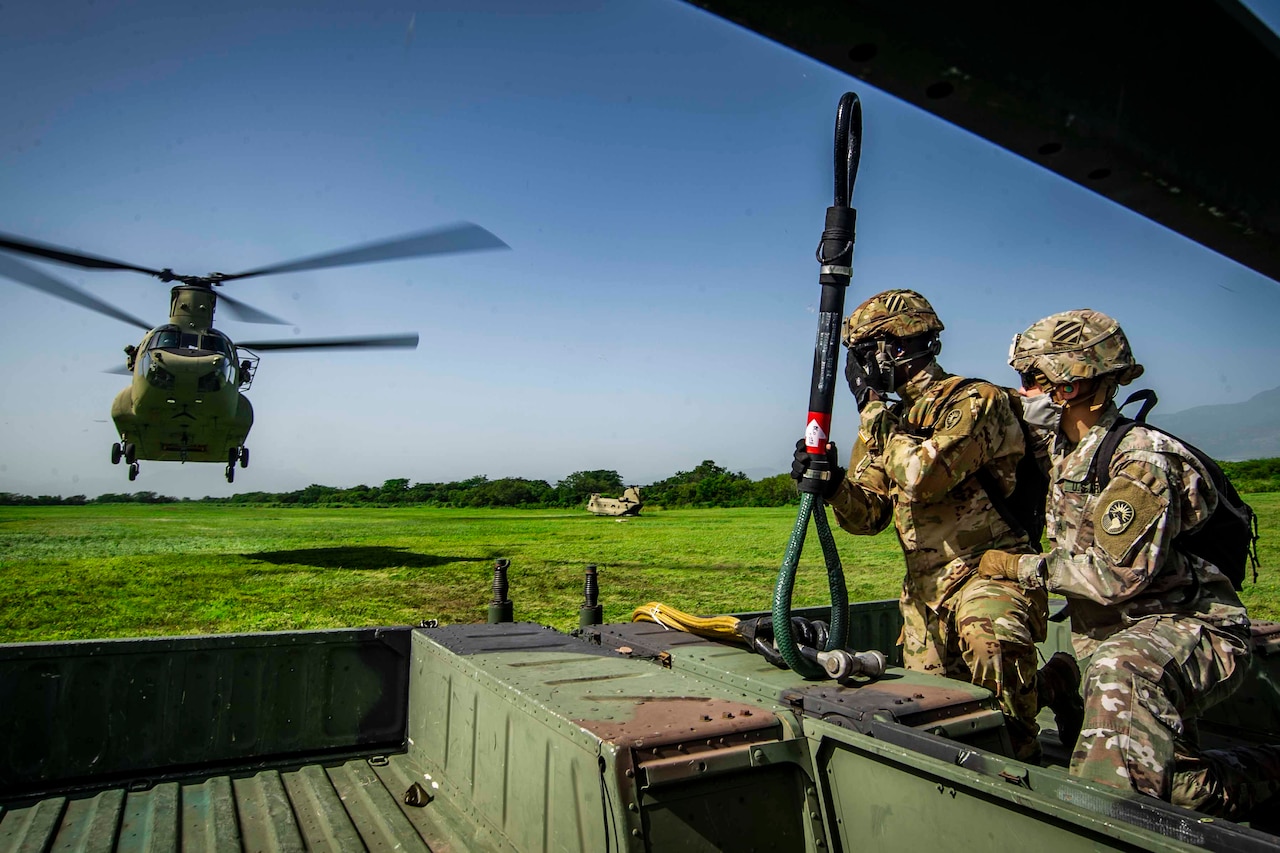 Two service members in a military vehicle watch as a helicopter approaches.