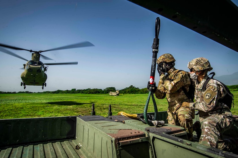 Two service members in a military vehicle watch as a helicopter approaches.
