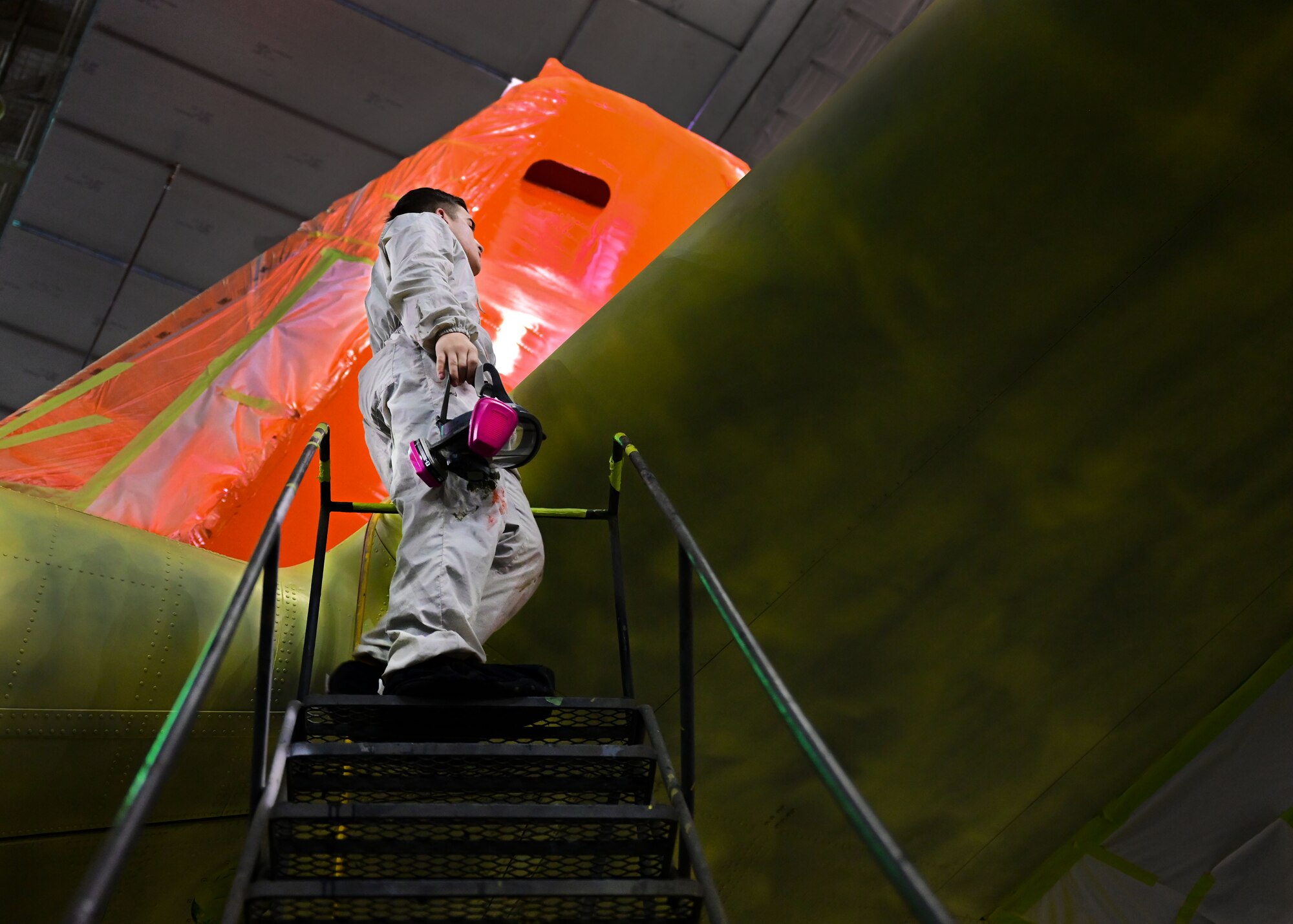 An Airman in a white suit speaks with another Airman on a set of stairs next to a C-130J.