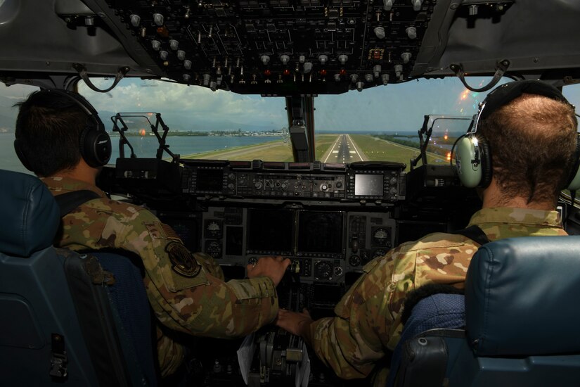 In a view from the back of a cockpit, two pilots look ahead at an airport runway.