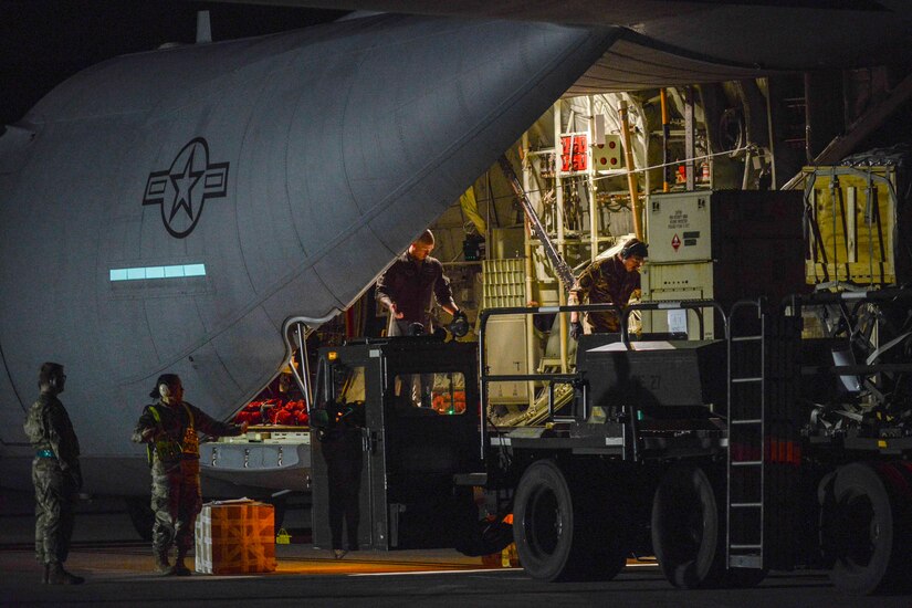 Two service members stand inside the cargo hold at the rear of an airplane, while one loads some pallets onto machinery. Two other service members stand outside the plane; a large box sits next to them.