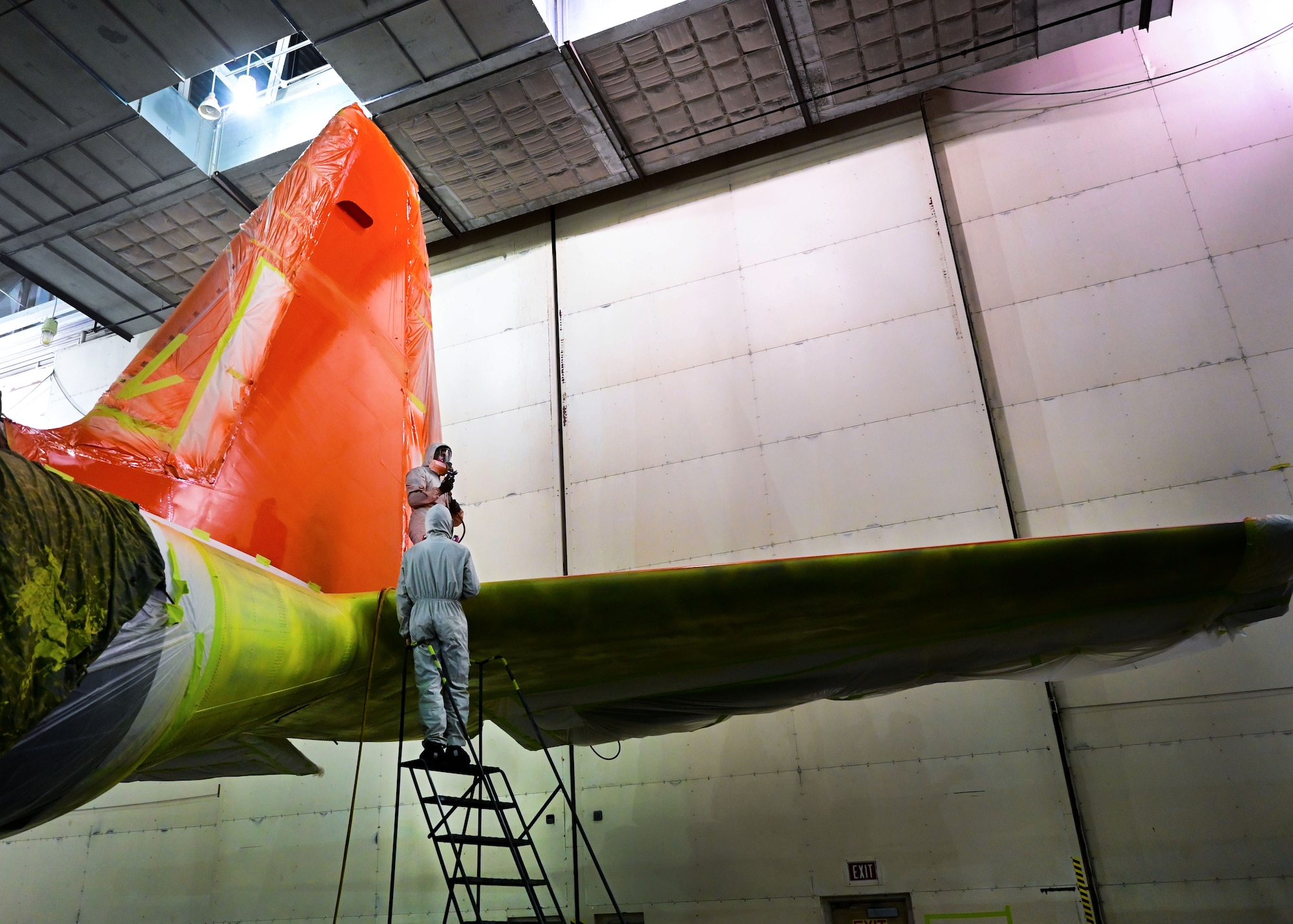 Two men in white suits prepare to spray bright orange paint onto the tail of a C-130J.