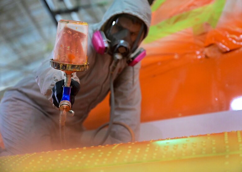 An Airman in a white suit sprays bright orange paint onto the tail of a C-130J.