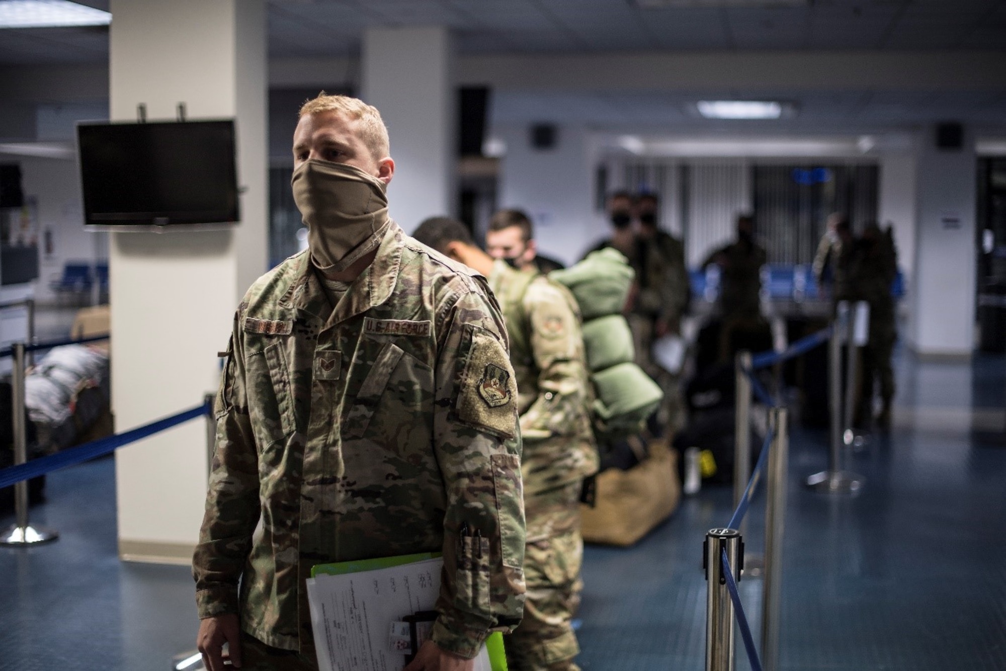 Men and women in military uniforms and face masks wait in a line inside airport-like surroundings