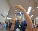 A service member in medical gear, a face mask and face shield uses a syringe