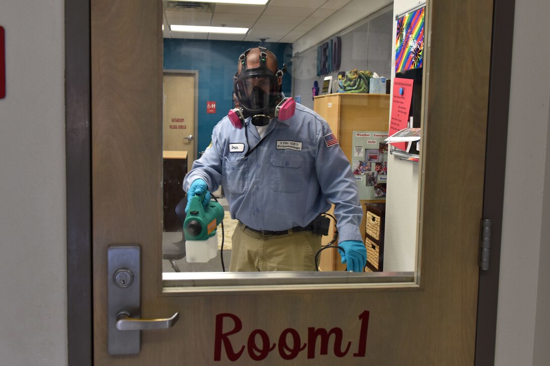 Behind a closed door with a glass window, a man dressed in a protective mask and gloves holds a machine with a canister attached to the bottom.