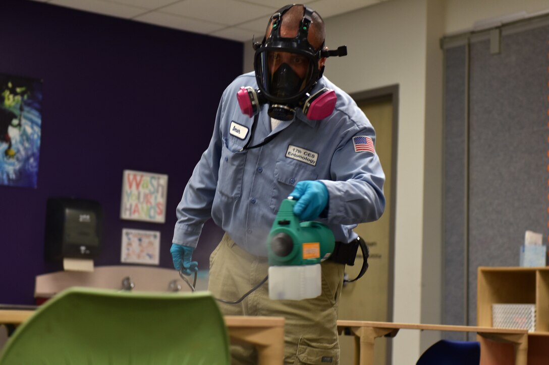 A man dressed in a protective mask and gloves sprays a mist onto a desk.