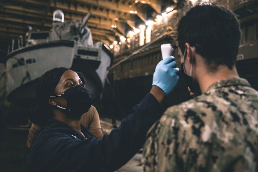 A Marine wearing a face mask and gloves takes the temperature of a Navy sailor wearing a face mask.