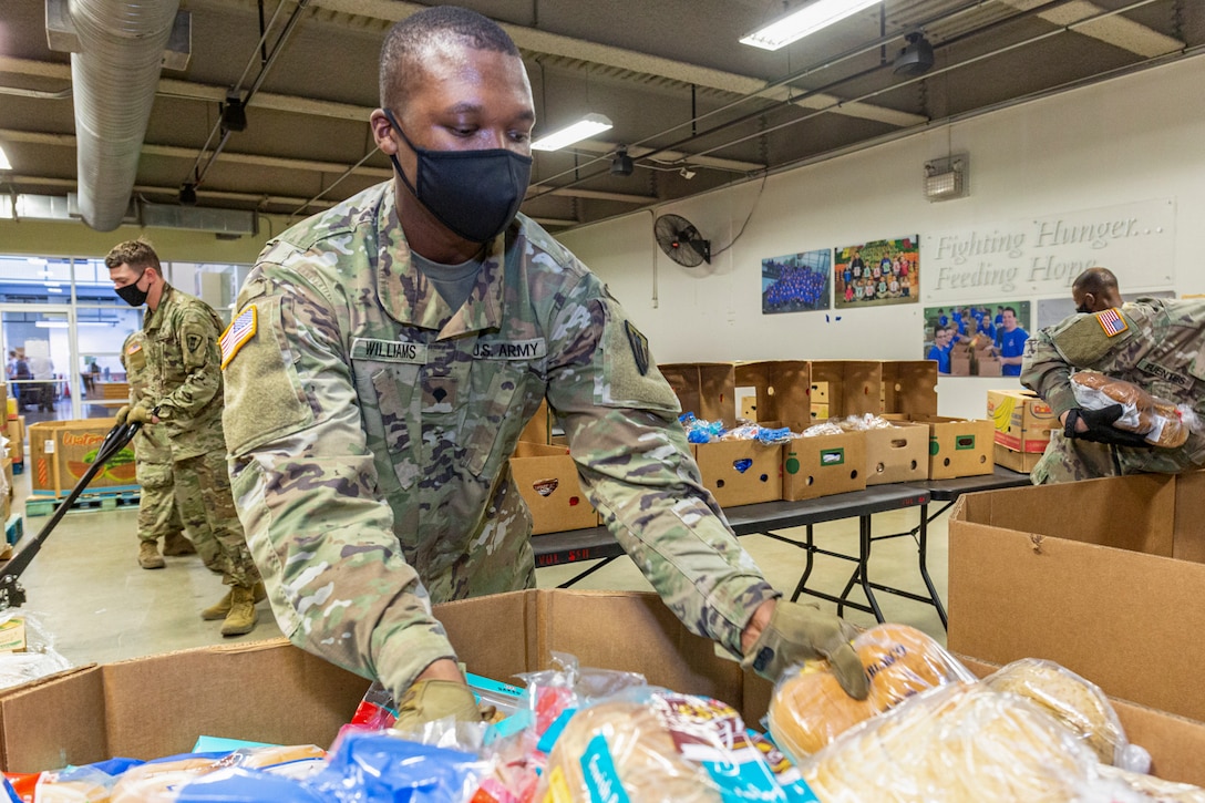 A male guardsman wearing a face mask and gloves helps distribute food.