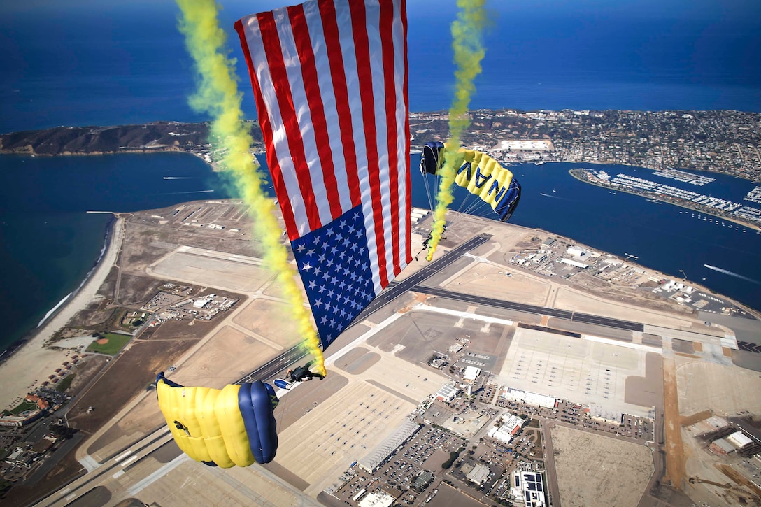 Sailors free fall while holding an American flag over a city.
