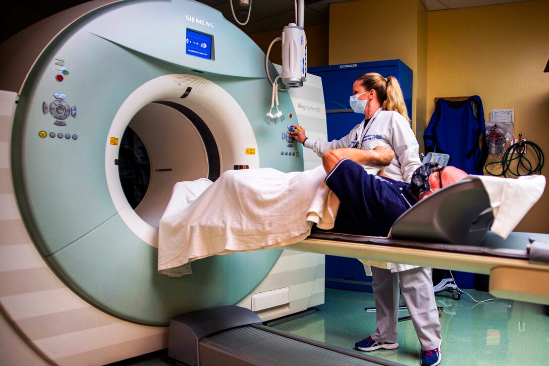 A woman wearing a face mask pushes some buttons on a large, medical imaging device as a male patient lying on a table prepares to enter the machine.