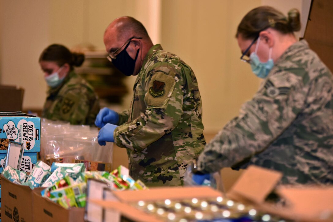 Two female and one male guardsmen, each wearing a face mask and gloves, put food into boxes.