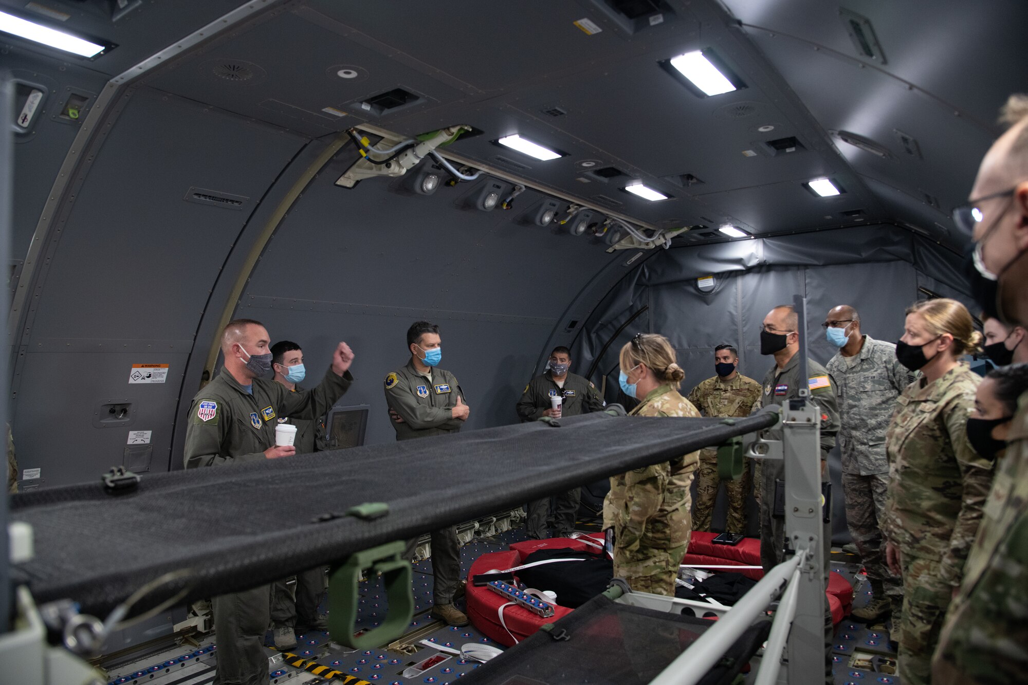 A group of people in Air Force uniform listen to three speakers in flight suits inside of a new cargo aircraft.