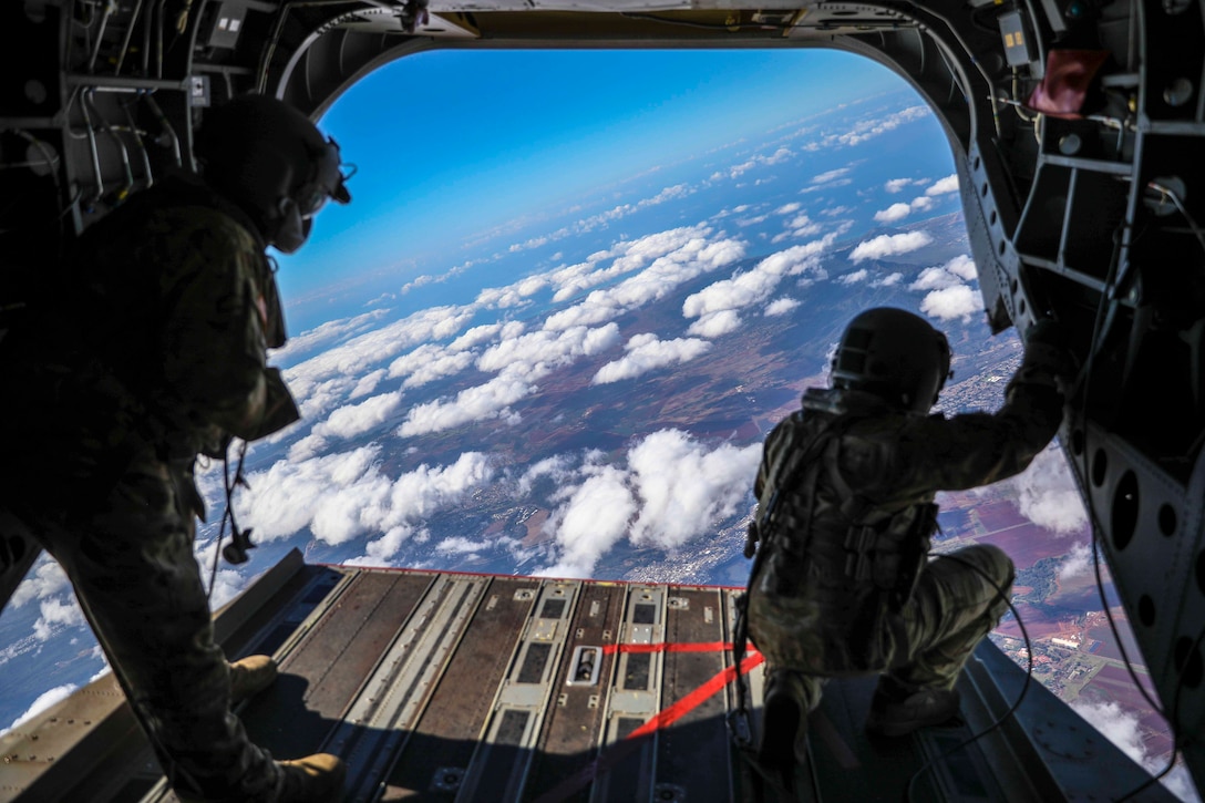 Two soldiers stand at the back of an aircraft; clouds seen below.