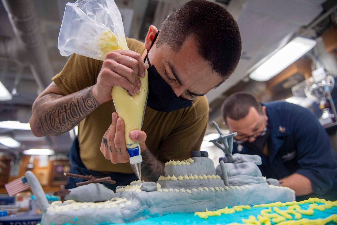 A sailor wearing a face mask uses a piping bag to decorate a cake.