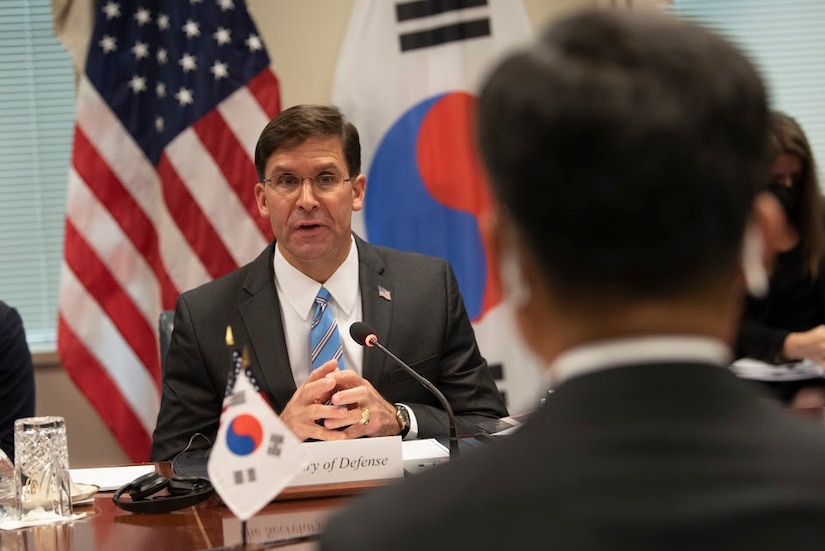 Man seated at a table speaks to another man across from him. The American flag is in the background.