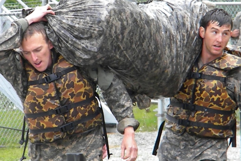 Two soaked soldiers in life vests carry a large camouflaged bag.