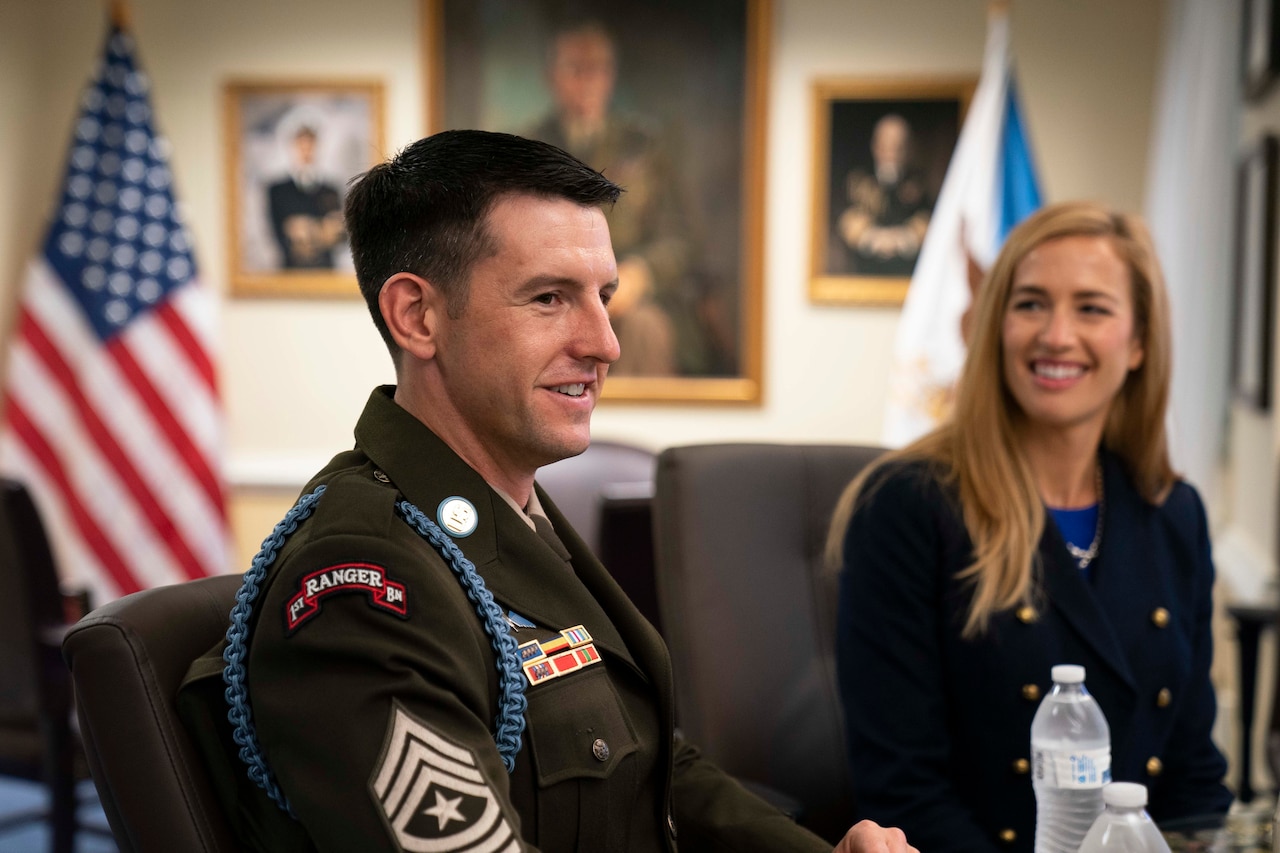 A man and woman, both smiling, sit in chairs in an office. An American flag is in the background.