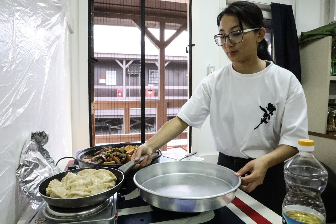 A woman stands over three pans cooking food on the stove.