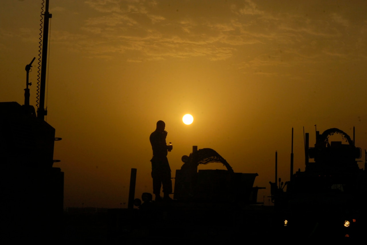 A man and military equipment are in silhouette against an orange, sunset sky.