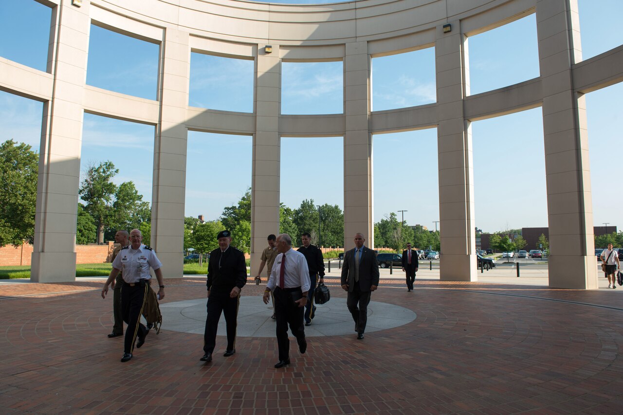 Multiple individuals, some in military uniform, walk outdoors through an array of columns.