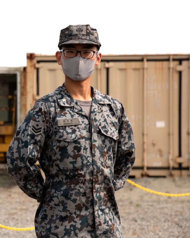 A male in uniform stand outside, looking directly at the camera with his hands behind his back