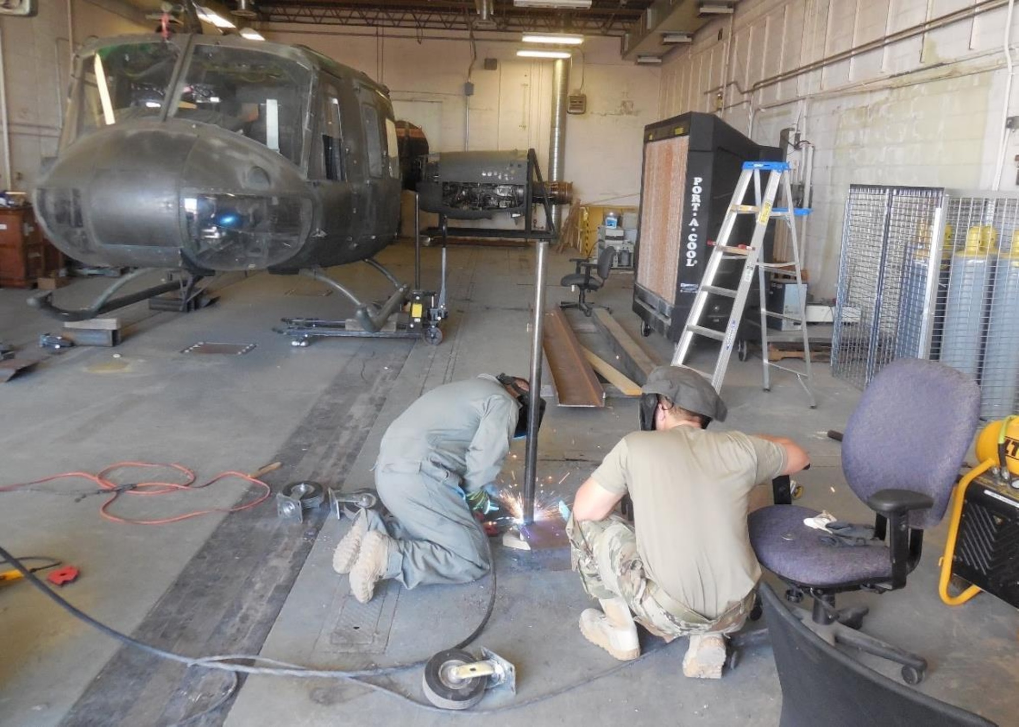A member of the 210th RED HORSE team (left) welds a wheel caster in July while another member looks on. The wheel caster aided in the transport of the UH-1 helicopter airframe pictured in the background. The airframe was relocated to the DNWS training site on base to be used as a training aid. Members spent a couple of weeks fabricating the casters provided by DNWS and rotated throughout the project to acquire metal cutting, welding and fabrication skills. (Photo by Matt Thompson)