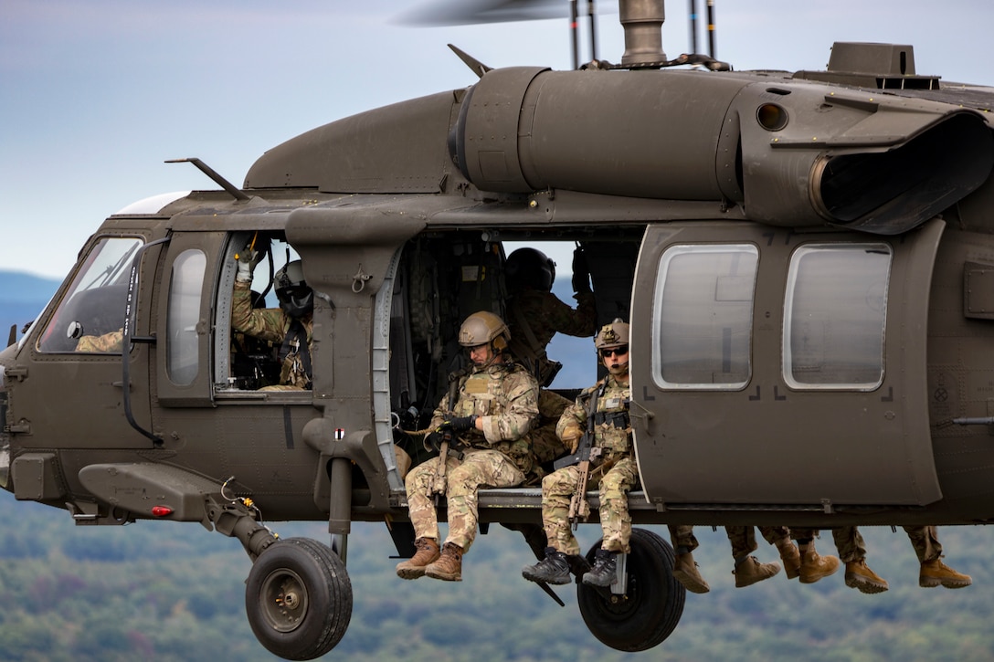 National Guard soldiers sit on the floor of a helicopter in flight and let their legs hang out from the aircraft.