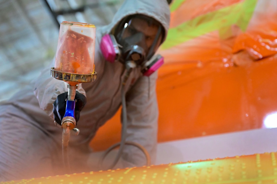 An airman in a protective mask and coveralls spray paints a metal object