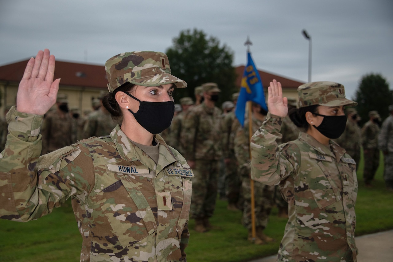 Two Space Force members wearing masks raise their right hands in front of a group of airmen on a parade-type field.